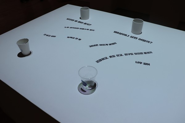 ▲본 전시의 4번째 공간 ‘카페100’의 카페 테이블에 질문과 답변이 나타나는 모습이다.